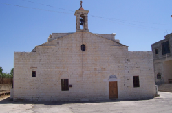 Front view of a beige stone church with a small bell tower and a wooden door under a circular window, on a sunny day